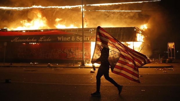 A protester carries the carries a U.S. flag upside, a sign of distress, next to a burning building Thursday, May 28, 2020, in Minneapolis. Protests over the death of George Floyd, a black man who died in police custody Monday, broke out in Minneapolis for a third straight night. (AP Photo/Julio Cortez)