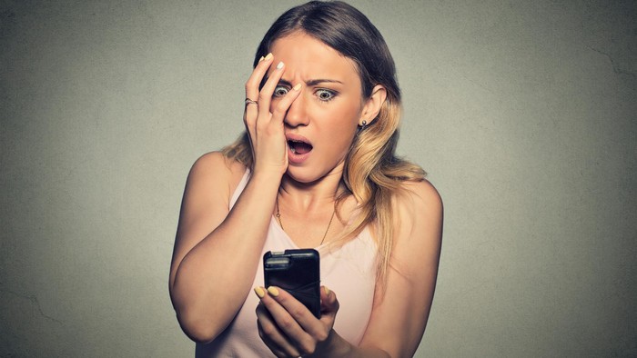 489896998 Closeup portrait anxious scared young girl looking at phone seeing bad news photos message with disgusting emotion on her face isolated on gray wall background. Human reaction, expression