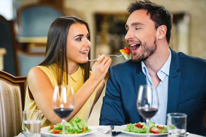 Photo of romantic dinner in expensive hotel. Young couple smiling while having dinner. Girl treating and feeding her boyfriend with salad