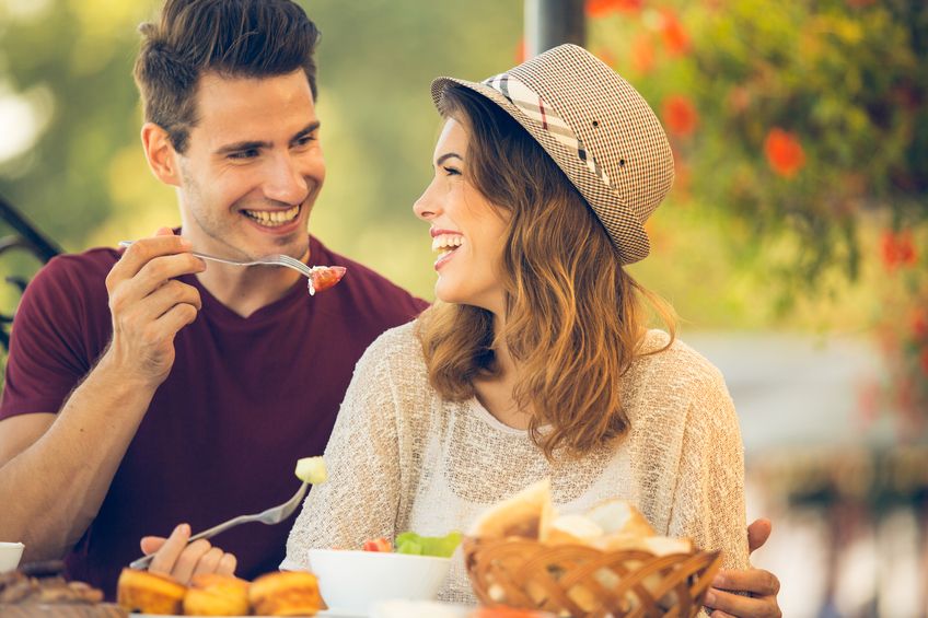 Photo of romantic dinner in expensive hotel. Young couple smiling while having dinner. Girl treating and feeding her boyfriend with salad