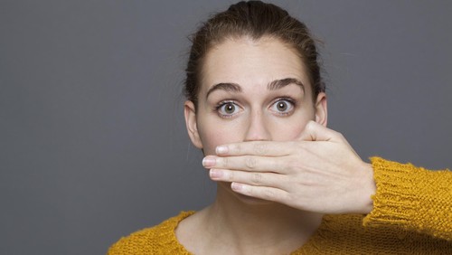 negative feelings concept - portrait of amazed beautiful 20s girl covering her mouth for gag metaphor,studio shot on gray background