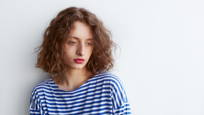 pretty sad hipster girl holding a skateboard on white background with copy space. Curly hairstyle young woman leaning against a wall thoughtful and looking down.