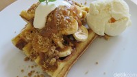 Waffle berbentuk persegi disajikan dengan irisan pisang dan disiram dengan saus karamel dan taburan biskuit. Ditambah dengan es krim vanilla. Nyam!