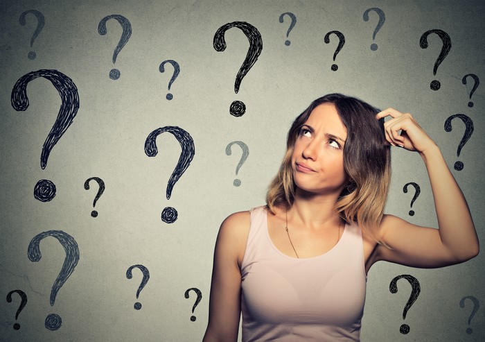 Thinking young woman looking up at many question marks isolated on gray wall background