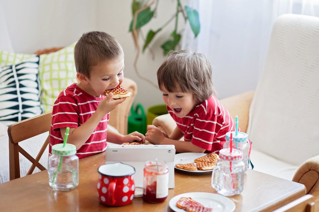 Two happy kids, two brothers, having healthy breakfast sitting at wooden table in sunny kitchen, eating waffles and watching cartoon on tablet