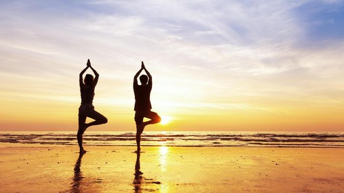 Two people practicing yoga tree position on the beach with beautiful sunset and reflection