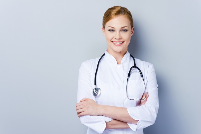 Cheerful female doctor in white uniform keeping arms crossed and smiling while standing against grey background