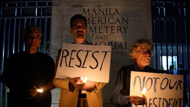 Omar Tiwana, Tim Ortiz, dan Donald Goertzen menyalakan lilin dan membawa poster memprotes pelantikan Trump. Mereka menggelar protes di luar pemakaman Amerika di kota Taguig, Manila.