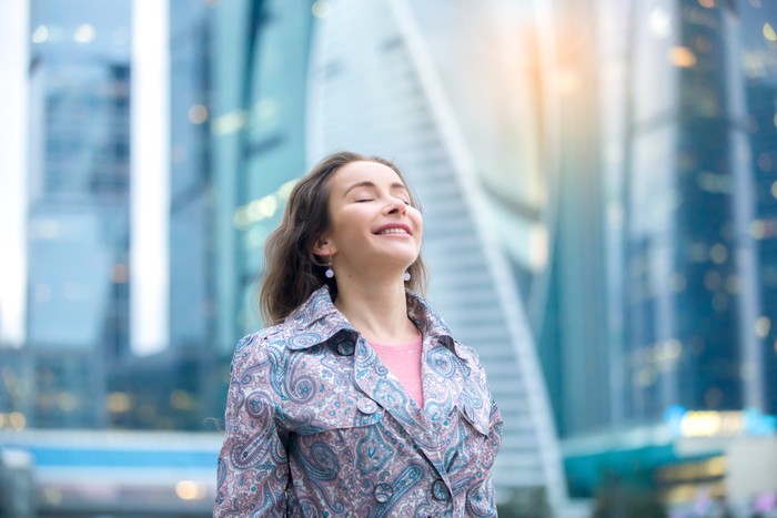 Portrait of young happy girl at street, her eyes closed with enjoyment, breathing at full outside. Lady feeling free, successful, ready to start day, visiting new town, blurred skyscraper background