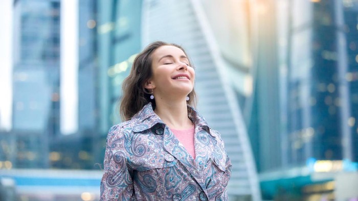 Portrait of young happy girl at street, her eyes closed with enjoyment, breathing at full outside. Lady feeling free, successful, ready to start day, visiting new town, blurred skyscraper background