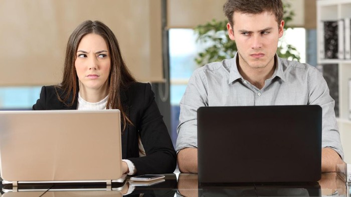Front view of two angry businesspeople using computers disputing at workplace and looking sideways each other with envy