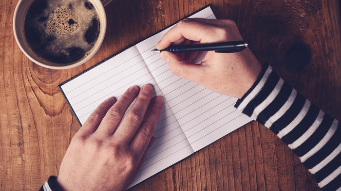 Woman drinking coffee and making a diary note, top view of female hands writing in notebook, retro toned image with selective focus.