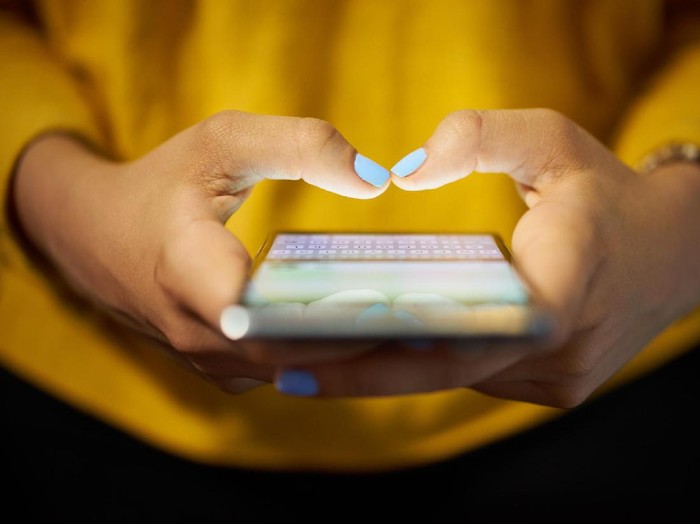 Young woman using cell phone to send text message on social network at night. Closeup of hands with computer laptop in background