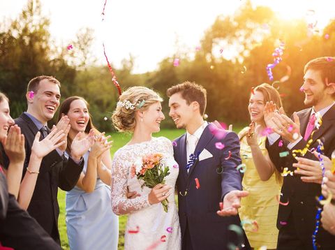 Full length portrait of newlywed couple and their friends at the wedding party showered with confetti in green sunny park