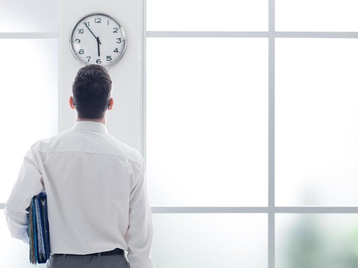 Businessman in the office staring at the clock, back view, time slave and stress concept