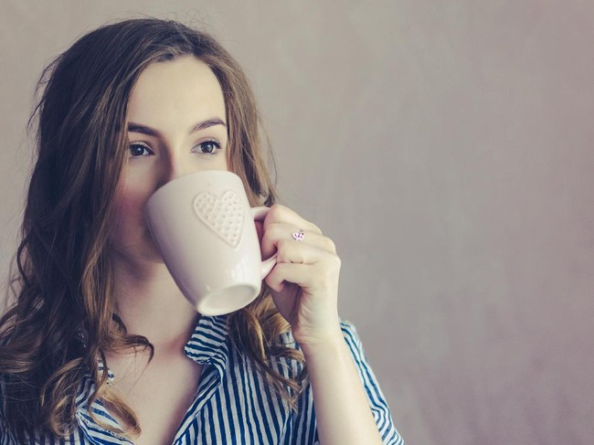 Beautiful teenage girl in striped shirt drinking coffee or tea from white cup in the morning. Portrait of sad young woman with curly fair hair wearing pajamas enjoying her beverage. Copy space.
