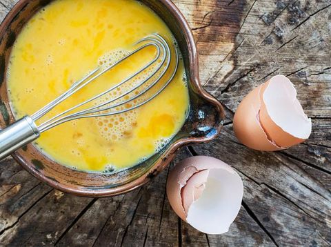 Beaten eggs and whisk in a ceramic bowl. In this photograph the bowl of eggs is placed on a rustic wooden surface, the whisk is placed in the bowl of eggs and is projecting out of the photograph. The egg shells from the beaten eggs are placed on the wooden surface. This photograph is taken with natural light. The image is intended for chefs, cooks, restaurants and egg sellers, as well as other.