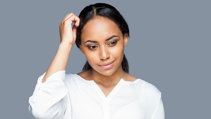 Thoughtful young African woman holding hand in hair and looking away while standing against grey background