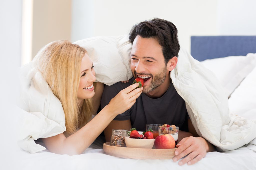 Portrait of a happy woman feeding strawberry to man for breakfast. Young smiling couple eating breakfast on bed. Happy young couple enjoying their breakfast at home.