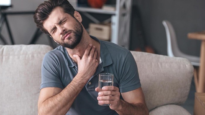 Frustrated handsome young man touching his neck and holding a glass of water while sitting on the couch at home