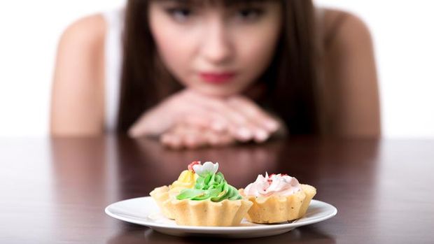 Young dieting woman sitting in front of plate with delicious cream tart cakes, looking at forbidden food with unhappy and hungry expression, studio, white background, isolated, close-up