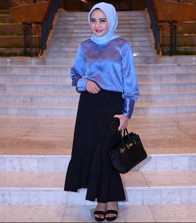 Gaya feminin Dewi Ramlan dalam balutan rok modifikasi warna hitam dan blouse biru satin. Gaya hijabnya tetap dibuat simpel hanya diikat ke belakang. Tas Hermes hitam memberikan kesan mewah dan elegan. Foto: Dok. Instagram @dewiramlan