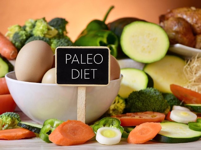 closeup of a signboard with the text paleo diet on a table full of different raw vegetables, a bowl with some chicken eggs and a chicken