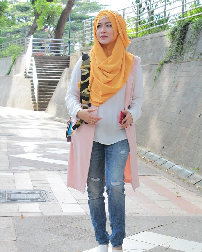 Kali ini penampilan Okie Agustina terlihat lebih kasual mengenakan celana jeans dan blouse putih. Ia menambahkan vest selutut yang membantu menutupi lekuk pinggang. Perpaduan kerudung kuning memberikan sentuhan eksentrik. Foto: Dok. Instagram @mrs.gdc13