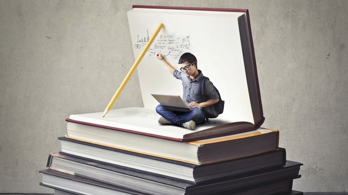 Young Asian guy with nerdy black glasses and backpack sitting in a huge book on top of a book hill writing some script on the page while looking at his laptop on his lap.