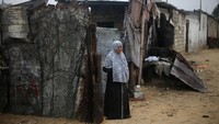 Seorang perempuan berdiri di depan rumahnya saat hujan deras di Raffah.