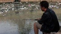 Seorang warga memancing di kali yang penuh sampah.