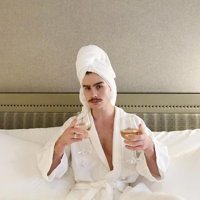 Pose seksi pria berkumis mengenakan handuk dan bathrobe di atas tempat tidur sambil memamerkan dua gelas wine di tangannya. Foto: Instagram