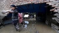 Seorang perempuan berdiri di samping rumah yang rusak karena banjir.