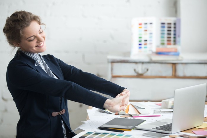 524148782 Portrait of young smiling woman wearing suit sitting at home office desk in front of laptop, stretching with enjoyment after the work is done, looking at screen with happy expression