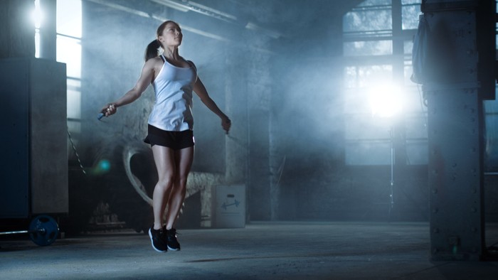 844485748 Athletic Beautiful Woman Exercises with Jump / Skipping Rope in a Gym. Shes Covered in Sweat from Her Intense Cross Fitness Training. Dark atmosphere.