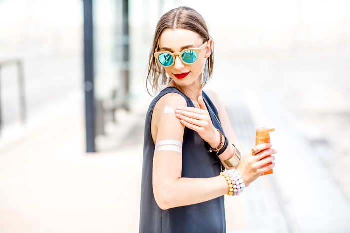 811467946 Young woman applying sunscreen lotion standing outdoors at the urban location during the sunny weather