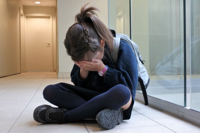 One young elementary school girl (age 7)  wearing school uniform and backpack sitting on a corridor floor crying. Childhood and education concept. copy space