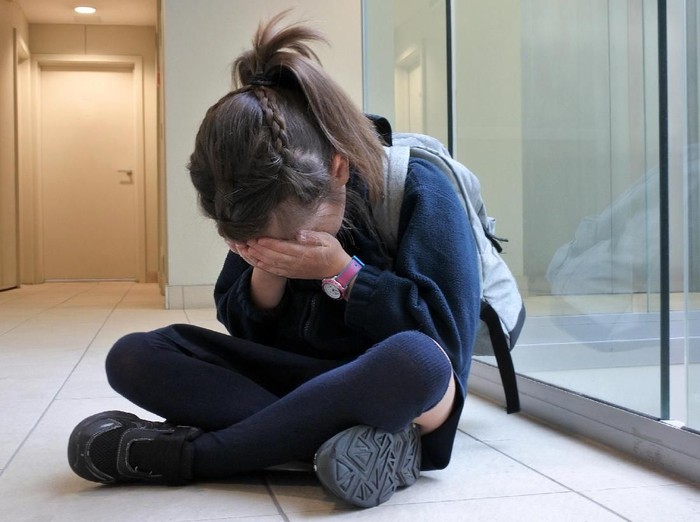 One young elementary school girl (age 7)  wearing school uniform and backpack sitting on a corridor floor crying. Childhood and education concept. copy space