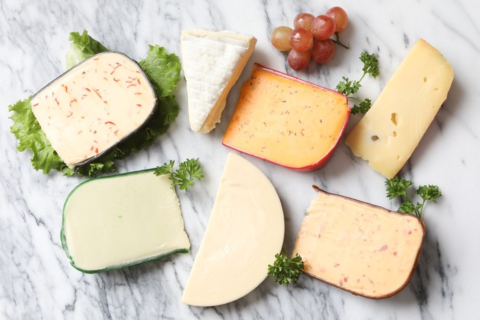 An overhead close up horizontal photograph of four artisanal and three traditional cheeses. Jalapeño peppadew,brie, buffalo wing flavor, Jarlsberg, smoked Gouda with bacon, sharp Provolone and Wasabi (horseradish) flavored cheese, garnished with curly parsley and some red grapes.