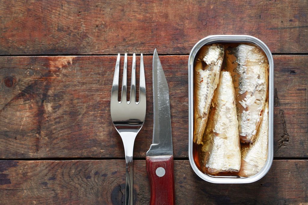 Open canned fish near fork and knife on wooden background with free space
