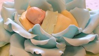 Tidak seperti kue, tapi lebih mirip seperti boneka kan? Fondant berwarna biru muda, hijau dan kuning ini dibentuk seperti bunga mawar yang sedang mekar dan seorang bayi yang sedang tertelap. Foto: Instagram @bakemeacakeorlando