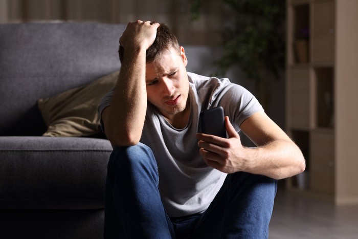 811258918 Single sad man checking mobile phone sitting on the floor in the living room at home with a dark background