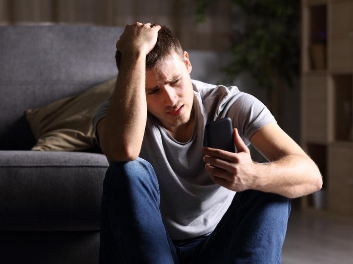 811258918 Single sad man checking mobile phone sitting on the floor in the living room at home with a dark background