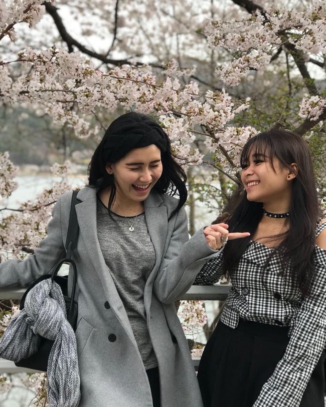 Tampil monokrom dengan kaus dan coat berwarna abu-abu, Ayu Ting Ting kembali menenteng tas Hermes seri Lindy saat berpose di antara bunga Sakura bersama adiknya, Syifa. Foto: Instagram