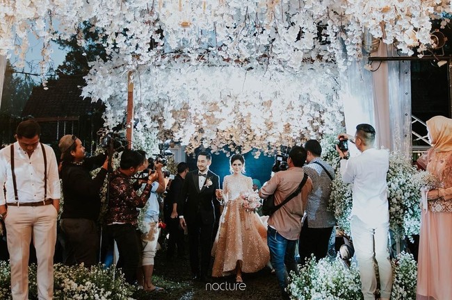Tak terasa nuansa hutan gelap dan menyeramkan ketika resepsi dihelat di malam hari. Jalan menuju pelaminan diberi sentuhan bunga putih di kanan dan kiri, pun langit-langit yang dibuat menggantung. Dekorasi serba putih yang elegan, mengingatkan akan dekorasi pernikahan Bella dan Edward Cullen di film Twilight. Foto: Dok. Instagram/Nocture