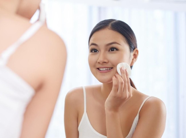 Smiling young woman applying toner on her face in front of mirror