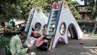 Seorang warga membaca koran di depan petugas yang tengah memperbaiki perosotan.