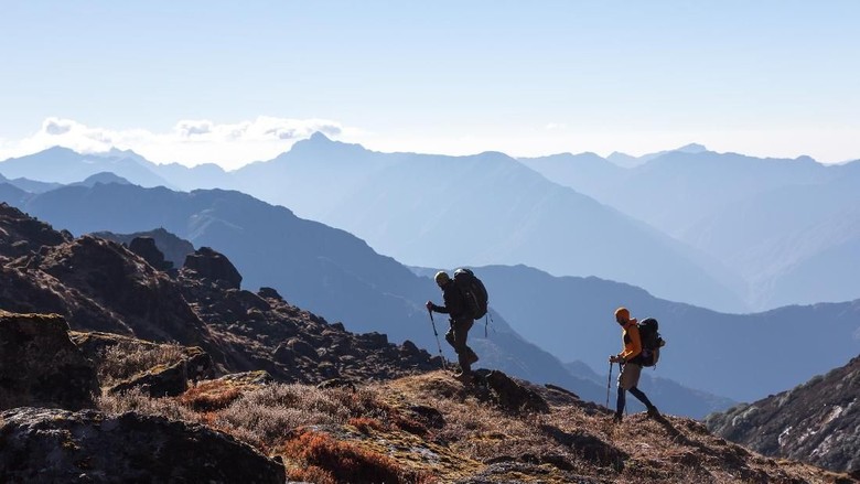 Two Men walking on rocky slope carrying Backpacks using trekking Sticks dressed in alpine Jackets and hiking Pants. Layered Mountains View beside Silhouetted of People