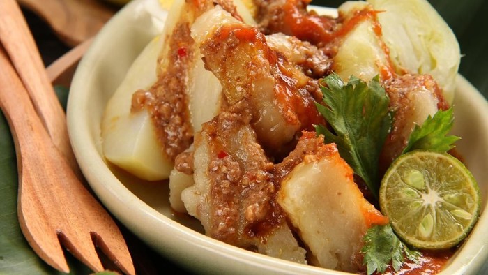 Siomay Bandung, popular Sundanese snack of steamed fish dumplings, potato, cabbage roll, and egg. Served with variety of condiments as spicy peanut sauce, chili sauce, and sweet soy sauce. A squeeze of lime is also added just before eating. This snack is plated on a oval-shaped ceramic plate; placed on a banana-leaf-lined wooden plate. Two small wooden forks are placed aside. This image was taken outdoor and used afternoon sun as the lighting source.