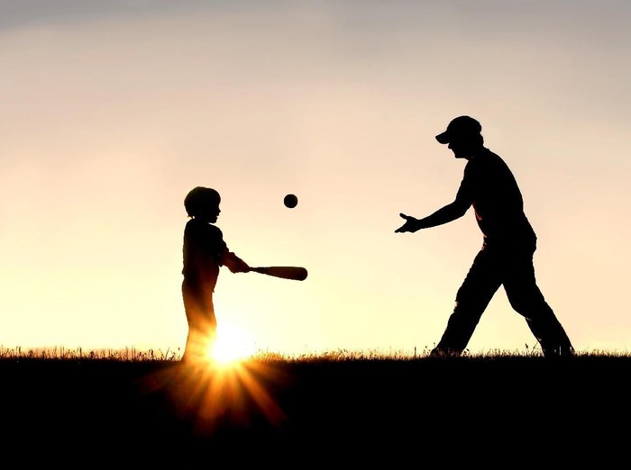 A silhouette of a father and his young child playing baseball outside, isolated against the sunsetting sky on a summer day.